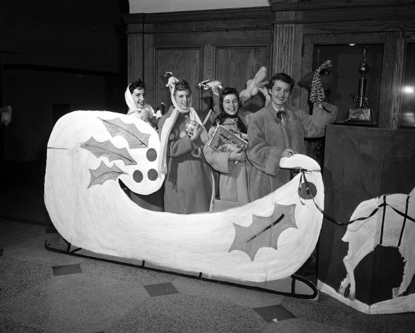 Four girls holding toys are standing behind a large cut-out Christmas sleigh at West High School. They are, from left: Betty Baird, Dagny Quisling, Mary Snell, and Gail Lawrenz.