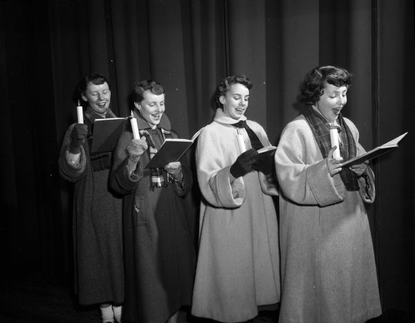 Four girls singing while looking down at their caroling books and holding candles and dressed for the outdoors. They are Diane Gensichen, Sally Tisdale, Susan Stauffer, and Joan Peck.