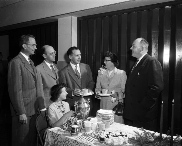 A welcoming reception honors Edmund Schwan, first principal at Monona Grove High School. Mrs. John Lyons sits at a table pouring tea, and standing from left are: E.L. Willett, R.K. Boutwell, Edmund Schwan, Mrs. Schwan, and Gerald Wilke.