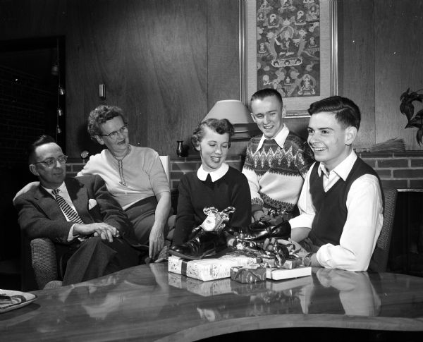 Jose Maria de Azcoitia of Barcelona, Spain (right) is shown with his host family at 6007 Winnequah Road. The host family consists of Mr. and Mrs. Allen Strang and their two children Betsy and Bill, students at Wisconsin High School. Jose is explaining Spanish Christmas customs, including January 6th "King's Day."