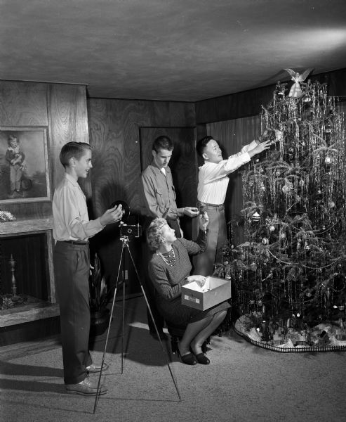 Hiroyoshi Yamamoto of Kurwe-shi, Japan (right) decorating a Christmas tree in the home of his hosts, the Dr. Frank K. Dean family. Tom (center) is standing behind his mother, Gladys, who is looking on with an ornament box on her lap. Jeff (left) is standing with a camera, flash, and tripod, ready to take a photograph.
