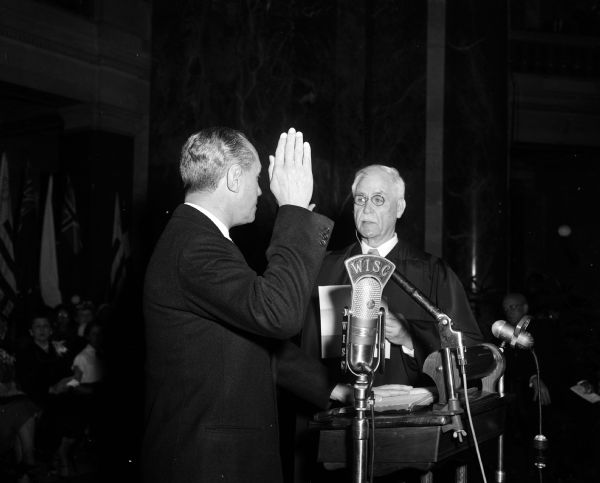 Governor Walter Kohler is shown with his right hand raised while taking his oath of office, being administered by Chief Justice Edward Fairchild of the Wisconsin Supreme Court. Governor Kohler is being sworn in for his third two-year term. In the foreground is a "WISC" radio microphone. An audience is looking on from the background.