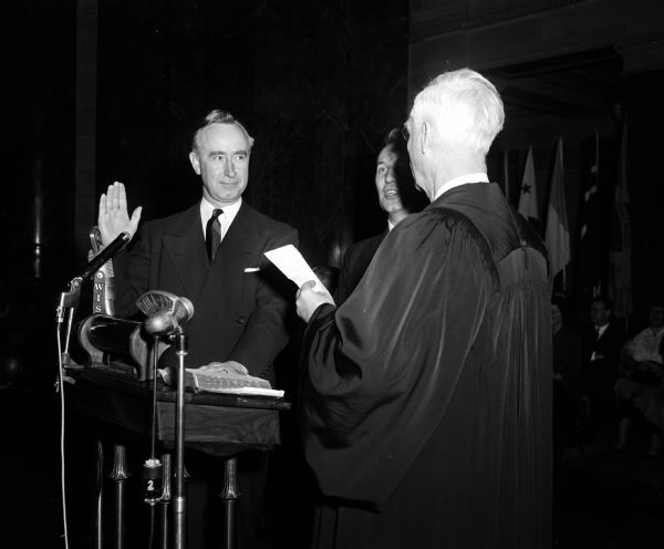 Attorney General Vernon Thomson, of Richland Center, is shown with his right hand raised while taking his oath of office, administered by a judge in a robe. Thomson is starting his third two-year term and he will later serve as governor (1957-1959). In the foreground is a microphone labeled "WHA-FM."
