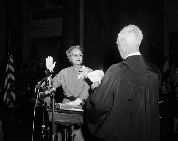 Glenn M. Wise (Mrs. John E. Wise) raises her hand while taking her oath of office before a robed judge. An audience looks on from the background. The original newspaper caption states: "Mrs. John E. Wise, Madison, becomes the first women to be sworn in for a state constitutional office."