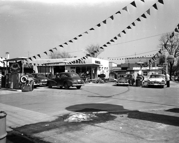 Exterior view of the Union Corners Shell Oil Station, 2601 East Washington Avenue at Milwaukee Street, viewed from across Milwaukee Street. The view includes six cars and three sets of gas pumps, strung banners, and two neighboring businesses to the east. The gas station was the site of the Union House tavern until 1954.
