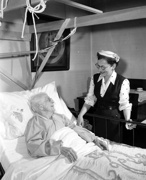 Sally Littig, chairman of the "loan closet" committee of the Women's Auxiliary of Dane County Medical Society visits with Nellie Kedzie Jones who lies in a loaned hospital bed after fracturing her hip.