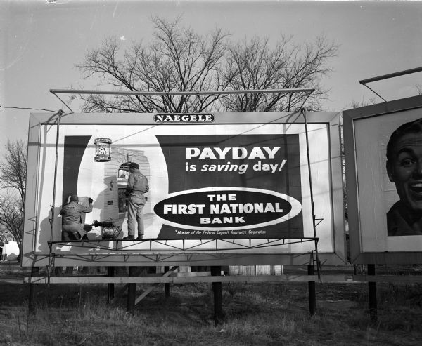 A two-man crew puts up a 24-sheet poster of outdoor advertising about the First National Bank. It takes 15 to 20 minutes to put the advertising up on this billboard.