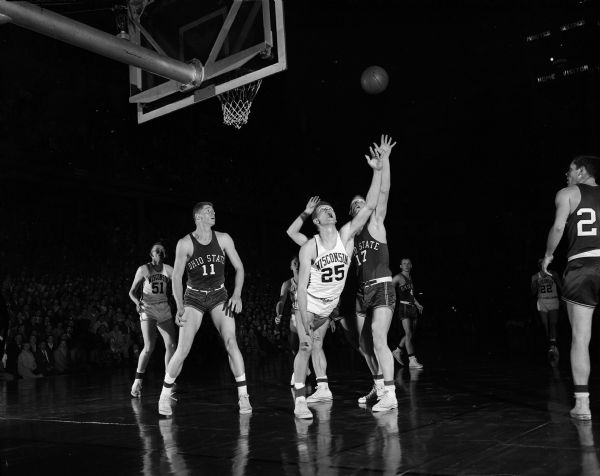"Curt Mueller (#25), who collected 15 points for Wisconsin, is shown as he put a little reverse 'English' on the ball and, believe it or not, scored on this fantastic shot in the first half. Ohio State players are Frank Howard (#11) and Jim Laughlin (#17)." Mueller is looking up with his hand in the air as the ball arcs up and behind him and eventually into the basket.