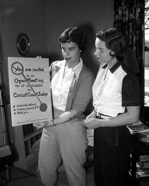 Margaret Johnson and Betty Hancock, members of the League of Women Voters, display a poster announcing a public meeting where candidates for mayor, 20th ward alderman, and supervisor will speak.