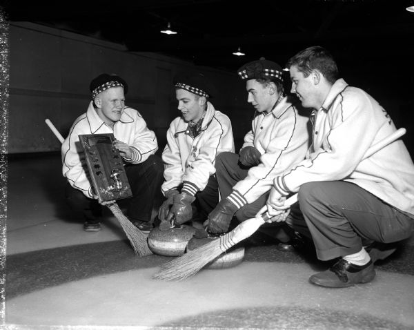 Four members of the Wauwatosa High School curling team wearing Scottish tams while squatting around two curling stones and two brooms and admiring their award plaque. The plaque reads "Lake Mendota Event Madison High School Bonspiel." The Wauwatosa Prep champions include, left to right, Skip Bob Brenckel, Mike Strock, Bob Fischer, and Jim See. They won the championship event in the Madison Invitational Bonspiel.