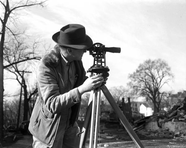 A man wearing a hat looks through the eye piece of his surveying instrument, called a 'theodolite,' which rests on a tripod.