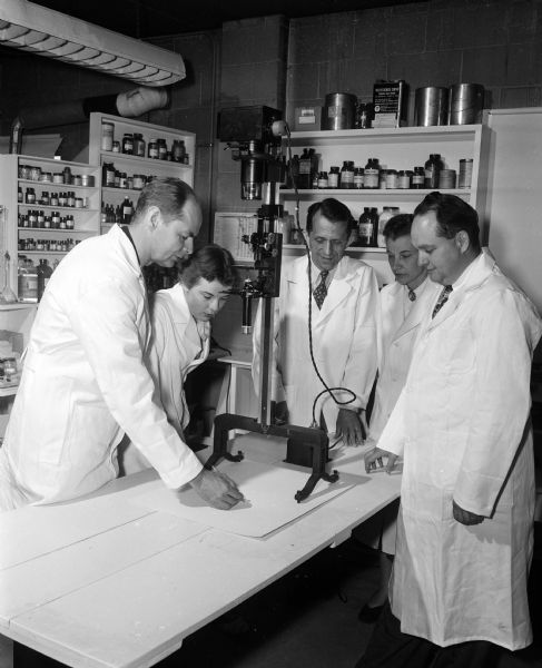 Five scientists wearing lab coats gather around an apparatus in a laboratory.