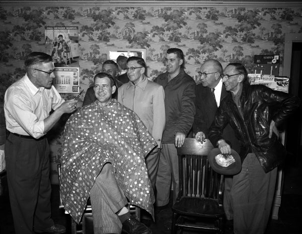 Barber Clarence (Hap) Lang discusses basketball with customer Joe Bernhard, while Nick Truckenbrodt, Herman Bings, Jerry Lochner, Ray Lueth, and Syl Fasking listen.