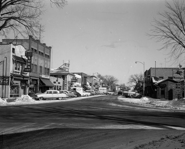 View of Water Street, looking north. Commercial buildings stand on either side of the street, including Lang's, Jack & Tony Superette, Ace Hardware, and Midway Stores at left and a Mobilgas station, Marty's Cafe and Bar, and Stratton's Footwear at right.