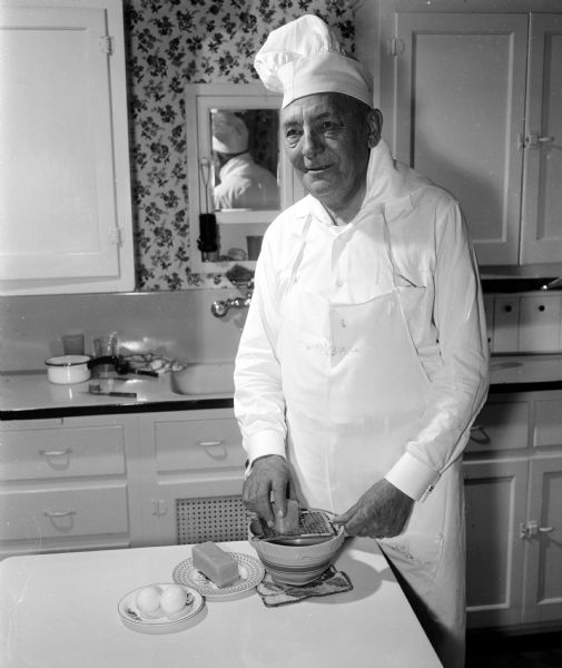 William H. Siemering stands in a kitchen grating cheese while wearing the traditional chef's apron, hat, and neckerchief. His specialty is "Eggs Wisconsin."