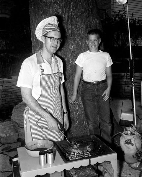 Jack L. Savidusky stands in his yard wearing a chef's hat and apron while barbecuing his specialty, "Mexican Barbecued Beef." His son Michael, 11, looks on.