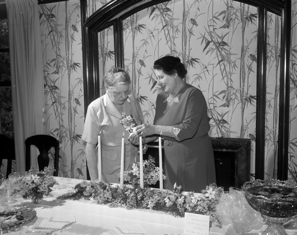 A floral arrangement typical of one used for a garden party in Holland stands on display at the YWCA benefit tea for internationally speaking University of Wisconsin foreign students. The tea took place at the official residence of University President, Edwin B. Fred and his wife, Rosa. At left is Rosa, hostess for the tea and right, Annie Van Der Stok, who arranged the floral centerpiece.