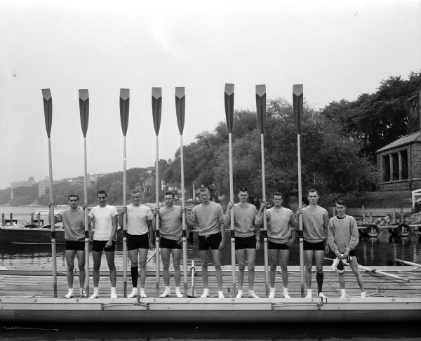 Nine men from the University of California rowing crew are holding eight oars while standing on a dock on the University of Wisconsin campus. Posing in the order in which they sat in the racing boat (scull), left to right, are: Dick Howard; Tom Grady; Dick Dobbins; Joe Fournier; John Peterson; John Dieterich; Gary Weyand; Gordon Raub; and Gene Hessel. The race was over a mile and three-quarters course on Lake Mendota.