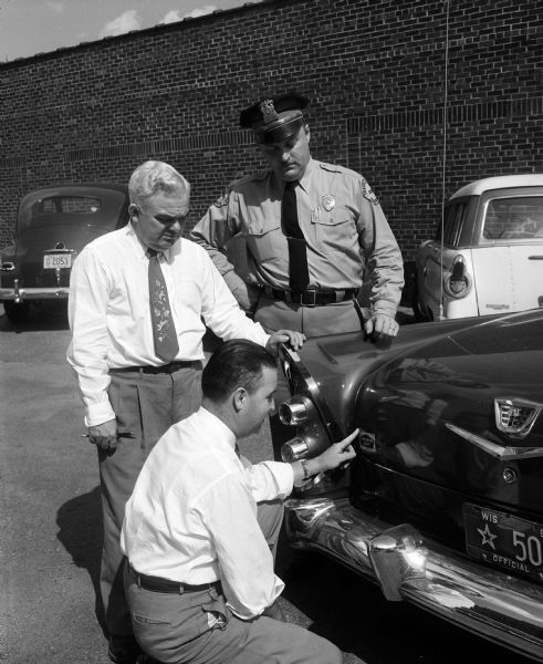 Two men look on as a third man crouches to apply a Goodrich Tire sticker to the back of a (Ford) police car. One of the men looking on is wearing a police uniform and visor cap with a metal badge. Two other cars are parked in the background.