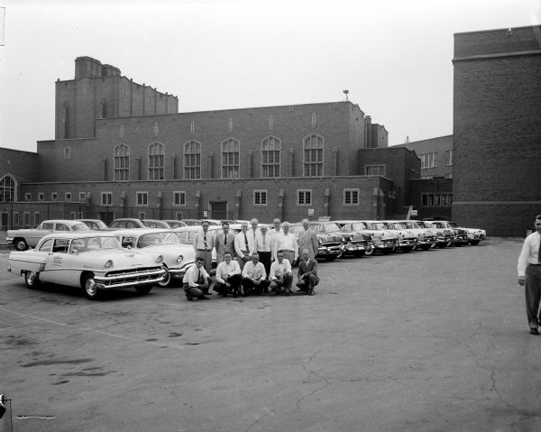 Twelve men are posing in front of over twenty similar automobiles parked in two neat lines on pavement in back of East High School. The men are auto dealers who have loaned cars or leaders of the drivers education program, possibly American Automobile Association (AAA) coordinators.