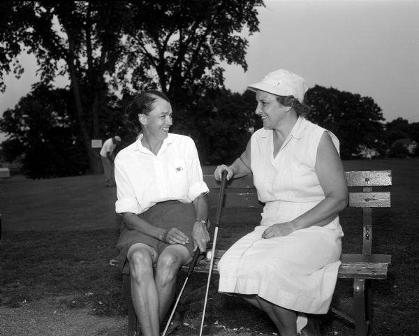 Alice Keller and Mr. F.G.H. Maloney converse while seated on an outdoor bench at Maple Bluff Country Club. Another golfer practices putting in the background.