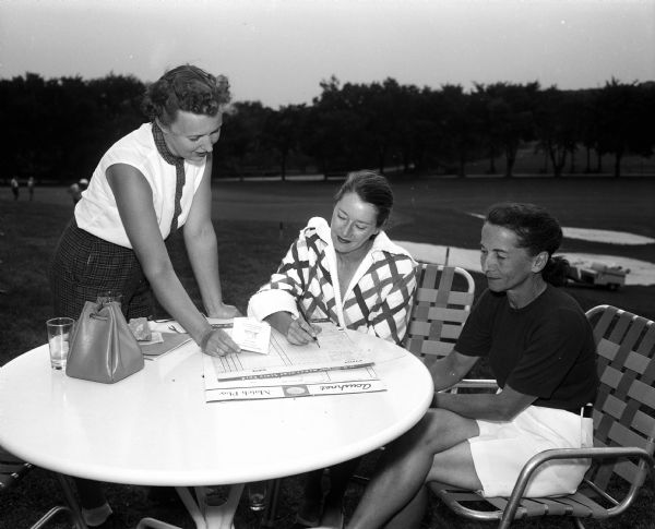 Jane Savidusky (left), Florence Lunger, and Jean Johnson check their scores after participating in a women's golf contest on Stagette Day. A golf fairway and other golfers are in the background.