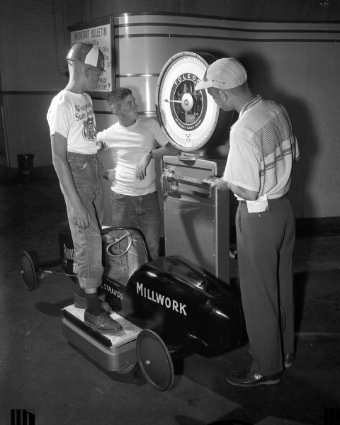 Alan Strauss stands with his derby car on the scale while Phil Peckham (left) records the weight; the boy and car cannot weigh more than 250 pounds. At right is Larry Jacobson, the 1953 champion.