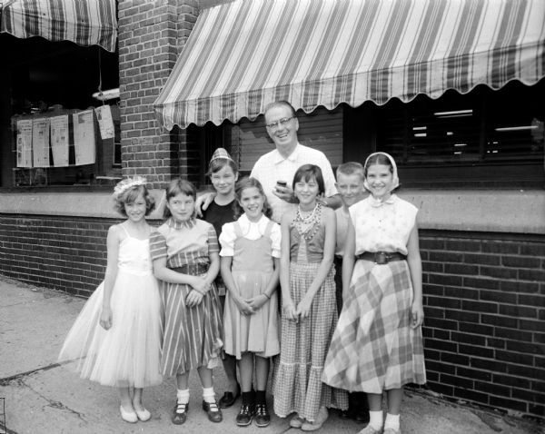 Six children and an adult posing in front of the <i>Wisconsin State Journal</i> newspaper office at 115 South Carroll Street. The children had put on a neighborhood circus to benefit the Roundy's Fun Fund and were at the office to present a check to Joseph (Roundy) Coughlin, State Journal sports columnist. Some of the children are dressed in circus costumes. Front row, left to right: Sandra Lee, Sally Gilbert, Charlene Quamme, Delores Gilbert and Sandra Quamme. Back row: Sandra Gilbert, Roundy, and Terry Lee.