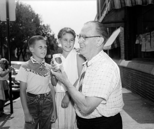 Two children and Roundy Coughlin posing outdoors in front of the <i>Wisconsin State Journal</i> office. Roundy is holding a can with the label "Donations for Roundy's Fun Fund." The children had earned the funds with a neighborhood puppet show. Right to left: Roundy, sports columnist for the State Journal; Frances Nelson and James Bucko.