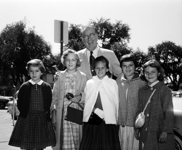 Outdoor group portrait of Joseph "Roundy" Coughlin, sportswriter for <i>Wisconsin State Journal</i>, with five girls who put on a neighborhood style show to raise money for Roundy's Fun Fund, a charity benefiting children with disabilities. Left to right: Linda Mosigin, Susan Werren, "Roundy," Mary Jo Sielaff, Sandra Stassi, and Nancy Werren.