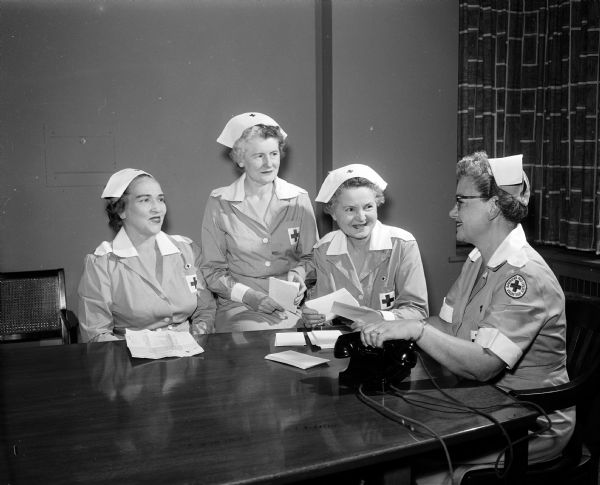 Four women wearing Red Cross Gray Lady uniforms, including nurse caps with Red Cross insignia, sit at a table and plan a united meeting of the four units of which they are chairmen. They are, left to right, Emily Harrington, Mendota State hospital unit; Evelyn Wheeler, Diagnostic Center; Iona Hein, county chairman; and Mrs. E. C.O. Erickson, Veteran's Administration hospital unit chairman.