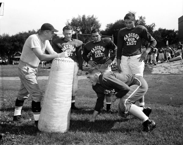 Madison East High School football coach Herbert (Butch) Mueller holds a dummy as player Dave McCloskey gets ready to charge during the first practice. Watching are Jon Rockstad, John Davidson, and Larry Davis.