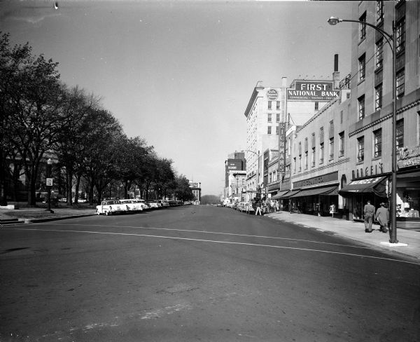 View of the South Pinckney Street side of the Capitol Square, one of four images making up a panorama of the southeast side of the Capitol Square. Pictured are the Belmont Hotel, the First National Bank, and other businesses on South Pinckney Street.