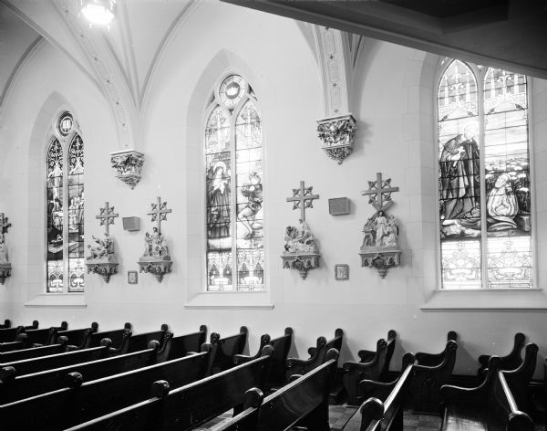 Interior view of St. Joseph's Catholic Church, Baraboo, decorated by Clemens Rath, showing side wall with stained glass windows and stations of the cross.