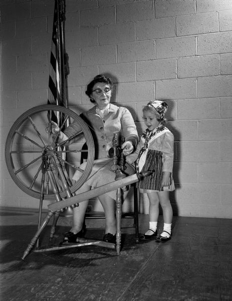 Anna Runstrom and her granddaughter, Erica Leigh Runstrom, demonstrate her spinning wheel at the East Side Businessman's hobby show. Erica is wearing a Swedish costume made by her grandmother.