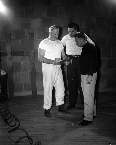 A gathering of three men, possibly University of Wisconsin boxing coaches, looking at a clipboard.