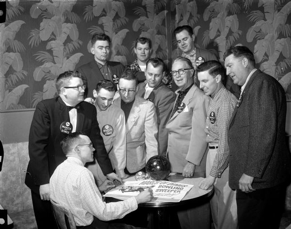 A committee plans the March of Dimes Bowling Sweepstakes. Seated at left is Chairman Dick Whalen. Standing front row left to right: Wayne Sweeney, Jim Wiedholz, Mike Whalen, Hi Yttri, Roundy Coughlin, Pete Viviani, Connie Schwoegler. In back, left to right:  Stan Mulrooney, Vic Comstock, Herman Ripp.