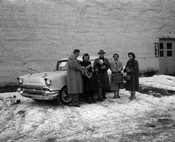 Five adults and three children posing in front of a prize new 1957 Chevorlet. On the left is Warner Young, chairman of the Madison Automobile Dealers Association prize committee, who presents the title to first place winner Mrs. Francis Hanson. To her left is her husband, and in front of them are their children, Lloyd, Paul, and Lynn. At right is second prize winner Agnes Rick, shown holding a portable television set, and Nancy Basford holding third prize, a transistor radio.