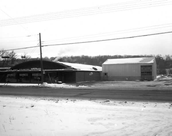 A metal store building and a metal warehouse building at 3320 University Avenue. In the background is a tree lined ridge. In the foreground is a two-lane road very near the buildings. The photograph was created to accompany an advertisement for a furniture sale.