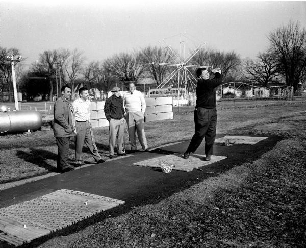 Four golfers with stand with their hands in their pockets while watching a fifth golfer, Frank (Moon) Molinaro power off a drive. The other four include, left to right: Steve Caravello, George Schiro, Bill Milward, and Bill Garrott. "Caravello is Wisconsin State Amateur champion and Milward is a former State Open champion."  Fairway Golf Practice Range was at 2205 South Park Street. Behind the five men in the background is the Fairway Amusement Park, a.k.a. "Kiddieland" at 2211 South Park Street. Behind the ferris wheel is a city bus.