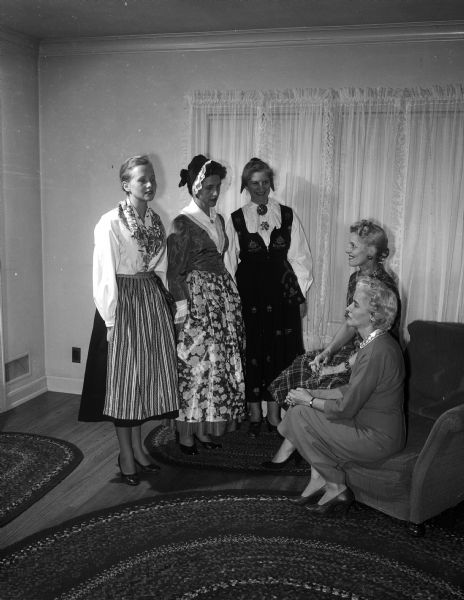 The original caption states: "Lynn Honeck, seated rear (right), wife of Wisconsin's new attorney general and a former professional model, is shown giving some suggestions to the faculty wives who will model foreign costumes for the University League's scholarship tea." Also seated in the right foreground is Ruth Liddle. Standing on the left are three faculty wives dressed in ethnic European clothes.Their names are, left to right: Jean Van Blaedel, Ruth Akert, and Eve Haugen.