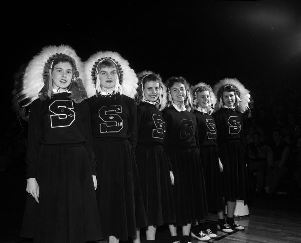 Six cheerleaders standing in a row. Each of the girls has a letter "S" sewn on the front of their sweaters and are wearing (Sioux) Native American "colorful" headdresses. Left to right are: Pat Otto, Judy Lorenz, Rosie Bosser, Jeri Sperberg, Nat Schroeder and Toni Brunner.  They are at the state boy's basketball tournament.
