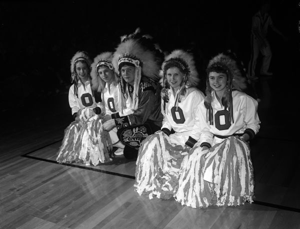 Four Osseo cheerleaders with the letter "O" sewed to their sweaters pose on one knee with pompoms covering their legs. They wear (Sioux) Native American headdresses while attending the state boy's basketball tournament. A man is posed squatting between the four girls, also wearing a (Sioux) Native American headdress. On his shoulders, cuffs, and pants are embroidered beadwork of floral designs in the Ho Chunk (Winnebago) Native American style.