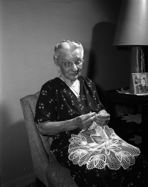Mrs. Friede (?) sits in a chair while crocheting a doily.