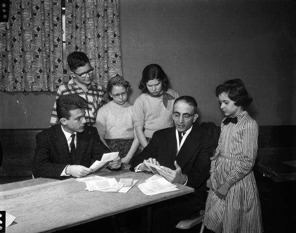 Danny Liarakos and Rev. Constantine Mager, priest of the new Greek Orthodox congregation, sitting at a table while reading letters. Four children are listening while gathered around them. The letters were written by Greek children thanking members of the Dane County Junior Red Cross for sending them gift boxes. The men are translating the letters which were written in Greek. The children, members of the Junior Red Cross, are, left to right: Bob McNown, Franklin School; Barbara Fausett, Sunnyside School; Mary Jordan, Franklin School and Loretta Tortorice, St. Joseph's School.