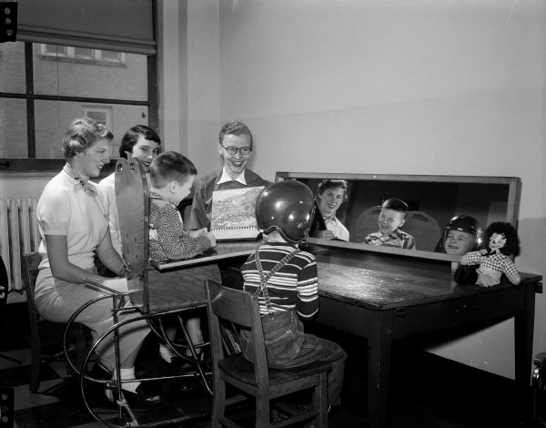 The caption states: "Alpha Chi girls majoring in child development, Gwyneth Fair, left, and Dorthy Bell, entertain two small victims of cerebral palsy. The little students of Mrs. Harriet Simpson, center, speech therapist in the Madison schools, are Donnie Pierce, 6, Lancaster, and Tommy Manning, 7, Richland Center, right, who attend the Madison Orthopedic school (inside Washington school, 545 W. Dayton st.)." Two college students and a teacher smilingly look on as two small students go about their activities while looking into a large mirror. One boy is harnessed to a large sled type chair and the other boy is wearing a football helmet.