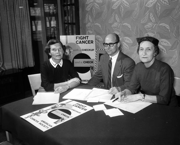 Two women and one man pose at a table with paper work. On the table and behind them are two identical posters, stating "Fight cancer with a checkup and a check; American Cancer Society."