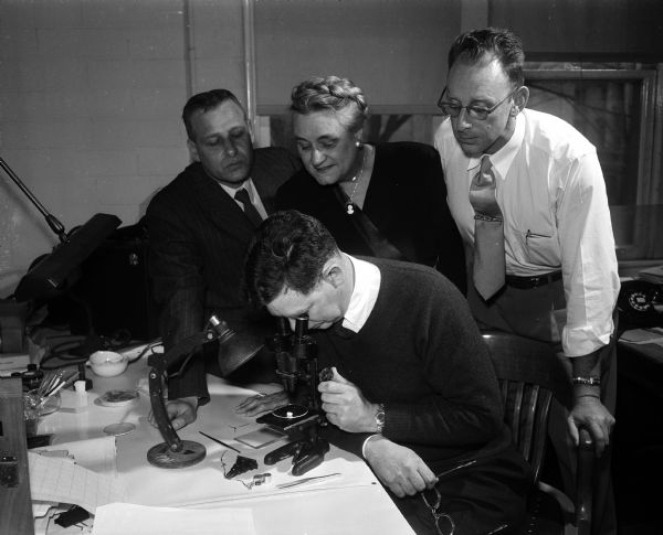 George Hanson (seated foreground center), state geologist, peers into a microscope while examining a diamond. Leaning in to look on are (left to right) Prof. Arthur Vierthaler; Agnes Hemingway, president of both the Madison Geological Society and the Madison Lapidary and Mineral Society; and Robert Black of the University of Wisconsin geology department.