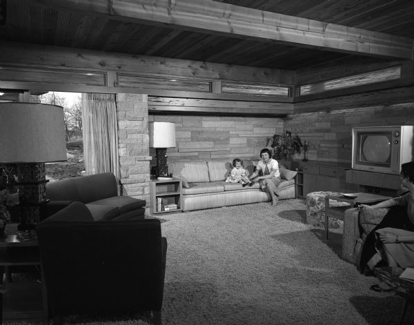 Joyce Bartell sitting on the sofa with her daughter Laurie, age three, in the living room of her and husband Gerald's home at 3938 Plymouth Circle in Madison. The room features Mankato stone, cedar ceiling beams, cypress panelling and gold textured rug and draperies. Another person is sitting on the far right.