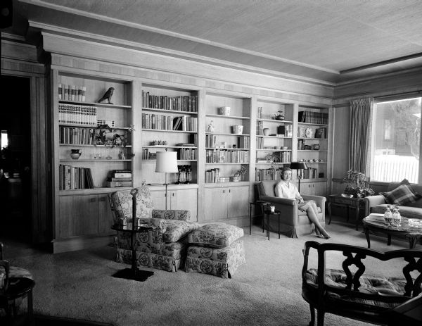 Marian Voight sitting in the living room of her home at 8 Fuller Court. The room features antique green grasscloth covering on the ceiling that matches the carpet, Limba wood paneling, and French Provincial furnishings.